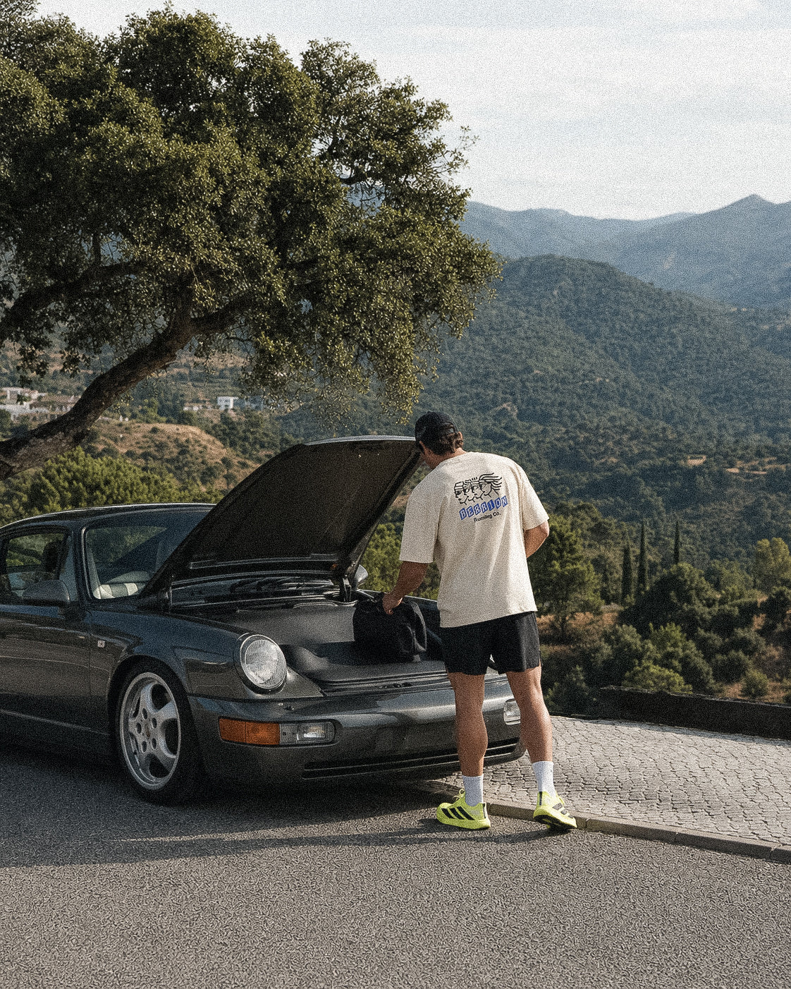 A man stands beside an open car hood on a paved road with mountains and trees in the background. He is wearing a cream graphic T-shirt, black shorts, white socks, and neon yellow-green sneakers, and appears to be interacting with a black bag near the car.